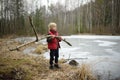 Little boy wearing red vest playing large branch on shore of forest lake on early spring day. Snow has just cleared  but surface Royalty Free Stock Photo