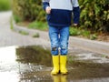 Little boy wearing yellow rubber boots jumping and having fun in puddle of water on rainy summer day in small town. Outdoors games Royalty Free Stock Photo