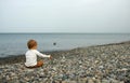Little boy sitting on stone beache and playing with stones. View from behind. Sea and cloudy sky on background Royalty Free Stock Photo