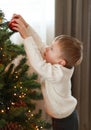 A little boy in a white sweater is diligently decorating the Christmas tree.