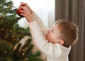 A little boy in a white sweater is diligently decorating the Christmas tree.