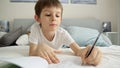 Little boy working on his homework in bed, writing in a notebook. Concepts of education, child development, and remote schooling Royalty Free Stock Photo