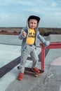 Little boy of 4-5 years old, in autumn summer in city on sports ground, learns to ride skateboard, smiles happy, shows