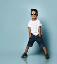 Little boy in sunglasses, white t-shirt, denim shorts and khaki sneakers. Smiling, put hands in pockets, posing on blue background Royalty Free Stock Photo