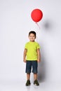 Little brunet boy in yellow t-shirt, denim shorts and khaki sneakers. Smiling, holding red balloon, posing isolated on white Royalty Free Stock Photo