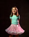 Little brunette child in blue t-shirt and poofy skirt. Smiling, hands folded behind her back, posing on black background. Close up