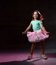 Little brunette child in blue t-shirt and poofy skirt. Smiling, hands folded behind her back, posing on black background. Close up