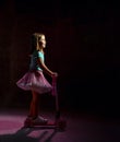 Little girl in blue t-shirt, poofy skirt and white sneakers. She is smiling and riding a scooter on black background. Full length