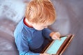 Little caucasian baby boy sits on the sofa using a tablet, touching screen. Red hair, casual wear, indoors, close up, copy space.