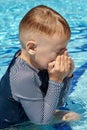 Little Caucasian boy with blond hair rubs his eyes with hands in summer swimming pool, or cries.