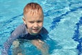 Little Caucasian boy with blond hair smiling in the summer swimming pool. Water drops on his face, wet T-shirt, blue color backgro Royalty Free Stock Photo
