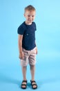 Little caucasian boy with blond hair stands on blue background. Suntan on hands and face, traces of mosquito bites on legs, summer