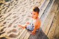 Little Caucasian boy child sitting on wooden pier sandy beach, summer time, sea vacation near water. The theme is the flow of time Royalty Free Stock Photo