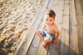 Little Caucasian boy child sitting on wooden pier sandy beach, summer time, sea vacation near water. The theme is the flow of time Royalty Free Stock Photo