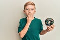 Little caucasian boy kid holding chocolate doughnut covering mouth with hand, shocked and afraid for mistake Royalty Free Stock Photo