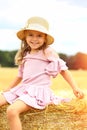 Little caucasian cute girl smiles in a wicker hat and a pink blue dress on a huge haystack on a mown wheat field. Agriculture. Royalty Free Stock Photo