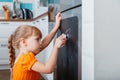 Little caucasian focused child girl draw with chalk on chalk board on refrigerator in kitchen at cozy home interior.