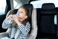 Little caucasian girl is driving in car, dreanking pure water from plastic kids bottle. Traveling on road in safe baby seats with