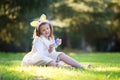 Little Caucasian girl in festive dress and bunny ears headband sitting on green grass and playing with Easter eggs Royalty Free Stock Photo