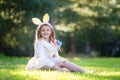 Little Caucasian girl in festive dress and bunny ears headband sitting on green grass and playing with Easter eggs Royalty Free Stock Photo