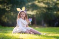 Little Caucasian girl in festive dress and bunny ears headband sitting on green grass and playing with Easter eggs Royalty Free Stock Photo