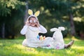 Little Caucasian girl in festive dress and bunny ears headband sitting on green grass and playing with Easter eggs and plush big b Royalty Free Stock Photo