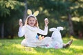 Little Caucasian girl in festive dress and bunny ears headband sitting on green grass and playing with Easter eggs and plush big b Royalty Free Stock Photo