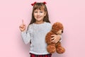 Little caucasian girl kid hugging teddy bear stuffed animal surprised with an idea or question pointing finger with happy face, Royalty Free Stock Photo