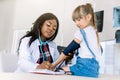 Little Caucasian girl at paediatrician office measuring blood pressure. Young African woman doctor measuring blood
