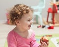 Little Caucasian girl in pink eats an apple and smiles an apple. Cheerful happy smiling baby with curly hair in the home interior Royalty Free Stock Photo