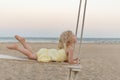 Little caucasian girl rides rope swing on sea background. Cute blonde toddler on the beach at sunset Royalty Free Stock Photo