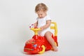 Little charming baby boy sitting on red and yellow tolocar, holding toy car in hands, wearing white body suit, has blond curly Royalty Free Stock Photo