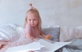 Little charming blond girl in pink dress holding a felt-pens and sitting on bed among paper. Royalty Free Stock Photo