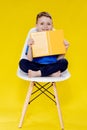 Little cheerful blond green-eyed boy 5-6 years old in a stylish blue T-shirt holding book and reading on yellow wall background, Royalty Free Stock Photo