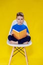 Little cheerful blond green-eyed boy 5-6 years old in a stylish blue T-shirt holding book and reading on yellow wall Royalty Free Stock Photo