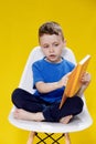 Little cheerful blond green-eyed boy 5-6 years old in a stylish blue T-shirt holding book and reading on yellow wall Royalty Free Stock Photo