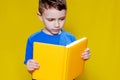 Little cheerful blond green-eyed boy 5-6 years old in a stylish blue T-shirt holding book and reading on yellow wall Royalty Free Stock Photo