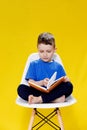 Little cheerful blond green-eyed boy 5-6 years old in a stylish blue T-shirt holding book and reading on yellow wall Royalty Free Stock Photo