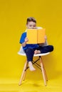 Little cheerful blond green-eyed boy 5-6 years old in a stylish blue T-shirt holding book and reading on yellow wall Royalty Free Stock Photo