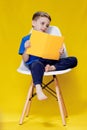 Little cheerful blond green-eyed boy 5-6 years old in a stylish blue T-shirt holding book and reading on yellow wall background, Royalty Free Stock Photo