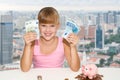 Little girl sitting on the desk with coins and pink piggy bank and holding in hands european currency over city