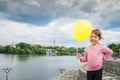 A little cheerful girl with a yellow balloon stands against the background of a river. A small child is in the city park. Royalty Free Stock Photo