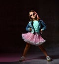 Little child in blue t-shirt, denim jacket, poofy skirt, sneakers, sunglasses. She smiling posing on black background. Full length