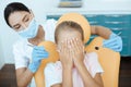 Little child covers face with hands, sits in medical chair with woman doctor holds instruments for examining patient Royalty Free Stock Photo