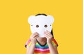 Little child girl holding blank white animal paper mask fronting her face isolated on yellow background. Idea and concept for kid