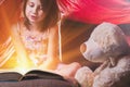 Little child girl reading magic book with teddy bear in tent before going to bed. Horizontal image Royalty Free Stock Photo