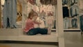 Little child, a girl, sits in a clothing store under the windows, dabbles and waits for her mother