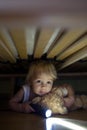 Little child, hiding under the bed, hugging teddy bear and holding flashlight Royalty Free Stock Photo