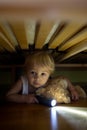 Little child, hiding under the bed, hugging teddy bear and holding flashlight Royalty Free Stock Photo