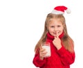 Little child in santa hat with glass of milk for Santa isolated. Beautiful little girl putting finger up to lips and ask silence Royalty Free Stock Photo